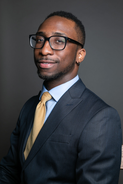 A portrait of Andrew Jones in a dark blue suit with a light yellow tie standing sideways looking at the camera.