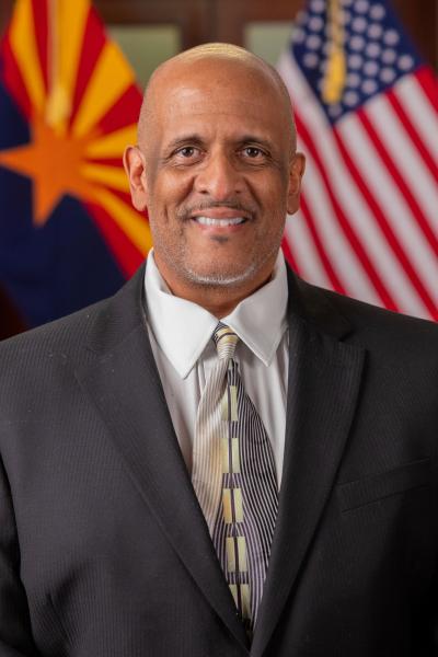 A portrait of Ken Owens Jr. in a suit with alternating American and Arizonan flags in the background.